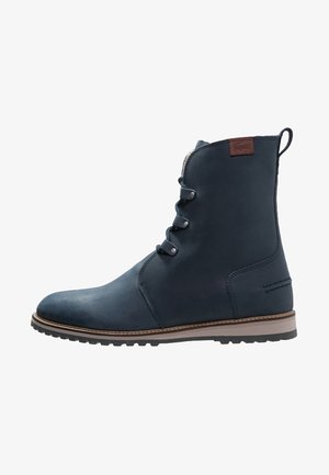 Bottine en cuir bleu marine avec trois œillets métalliques, semelle marron contrastante et patch logo marron près du haut à l'arrière.