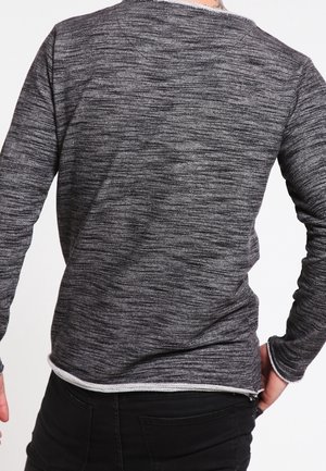 Mann, der einen langärmligen, dunkelgrauen, strukturierten Pullover und schwarze Hosen trägt, von hinten gezeigt, mit einer Hand in der Tasche.