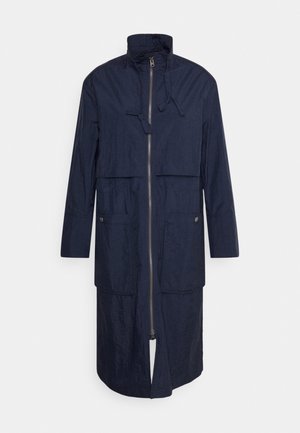 Tummansininen pitkä takki, joka on valmistettu teksturoidusta materiaalista, jossa on korkea kaulus, etuvetoketju, kaksi läppätaskua ja suora leikkaus.