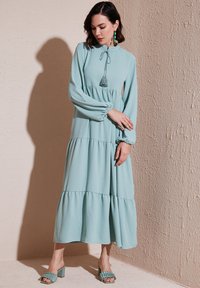 Light blue long-sleeve dress with a high neckline and tassel tie, featuring tiered ruffles. The fabric is soft, and flowy with a smooth texture.