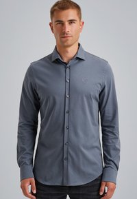 Joven con una camisa gris ajustada de manga larga y botones, de pie frente a un fondo liso de color gris claro.