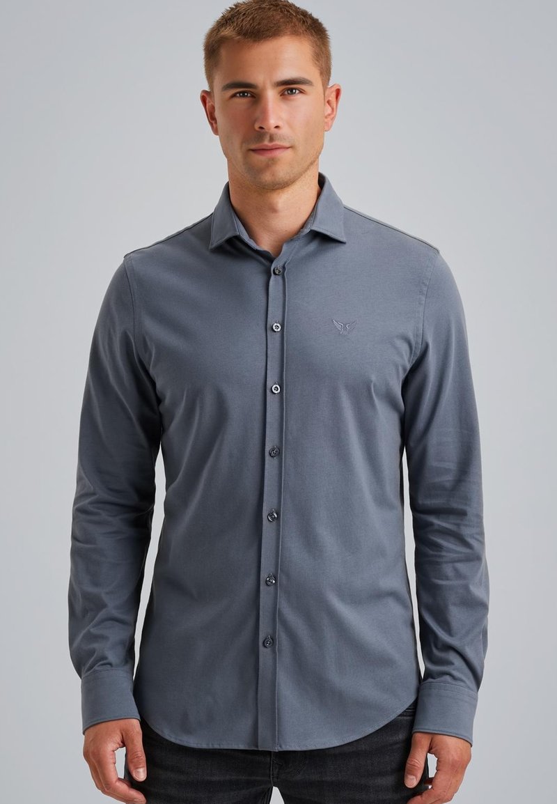 Joven con una camisa gris ajustada de manga larga y botones, de pie frente a un fondo liso de color gris claro.