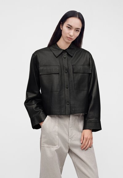 Femme portant une veste en cuir noire boutonnée avec des poches avant et un pantalon beige clair sur fond uni.