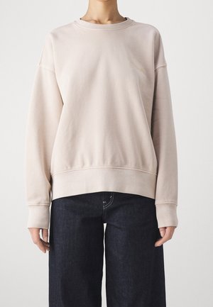 Persoon draagt een beige trui met ronde hals en subtiele borduursels op de borst, gecombineerd met donkerblauwe losse jeans.