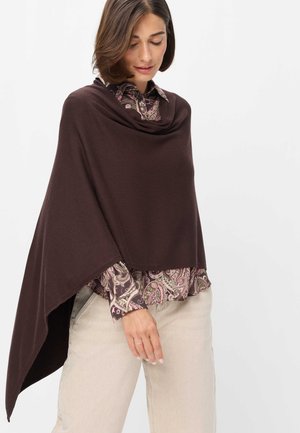 Kvinde iført en mørkebrun asymmetrisk poncho over en skjorte med paisleymønster og lyse bukser, ser ned med et blidt udtryk.