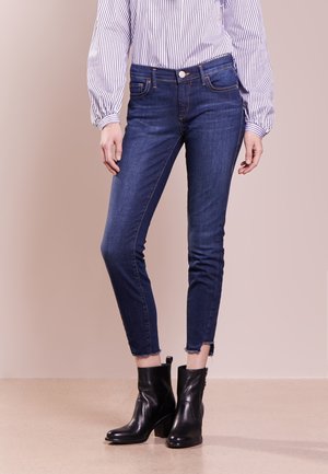 Femme portant un jean skinny bleu foncé avec des ourlets effilochés, des bottines noires et un chemisier blanc à rayures verticales noires.