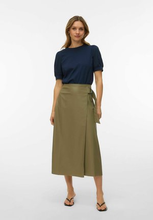 Woman wearing navy short-sleeve top and olive green wrap midi skirt with buckle, standing against plain white background.