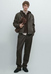 Bruine hoodie met rits, gedragen over een donker trui en gestreept overhemd, gecombineerd met bijpassende broek. Houdt een opgerolde deken vast.