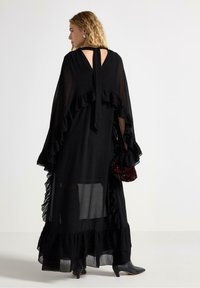 Svart genomskinlig klänning med volangdetaljer, öppen rygg och snördetalj. Har en lager-på-lager fåll och är kombinerad med en texturerad röd väska.