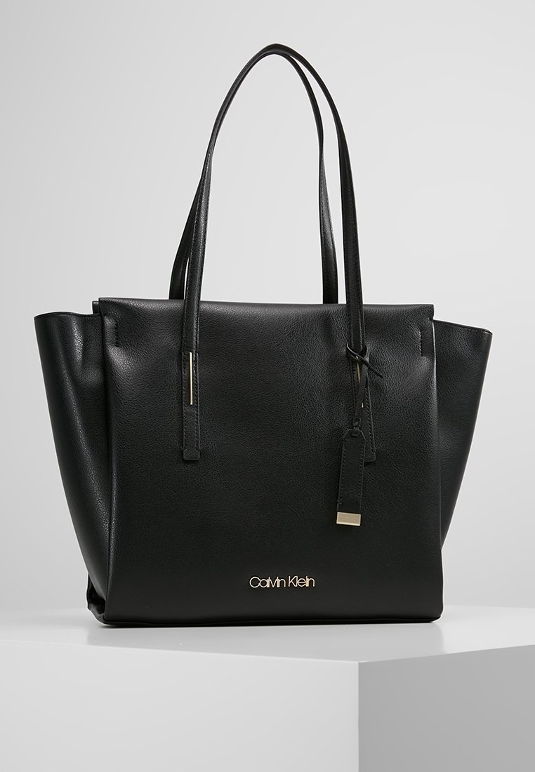 Black leather tote bag with two long handles, textured surface, gold hardware accents, and a logo on the front.
