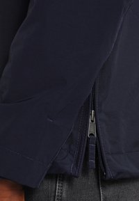 Marineblauwe jas met een gladde textuur, ritssluiting en contrasterende ritsdetails. De mouw heeft een strakke naadafwerking en een getailleerde pasvorm.