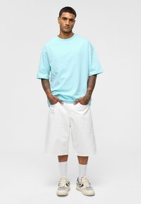 T-shirt oversize en coton bleu clair, shorts blancs arrivant au genou, chaussettes blanches et baskets avec des accents gris, se tenant devant un fond neutre.