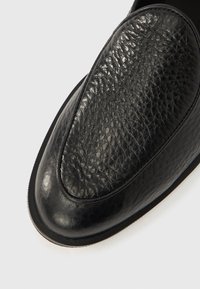 Chaussure en cuir noir avec une tige texturée et un bout lisse. Présente une forme arrondie et un design minimaliste. Semelle fine avec une couture légère.