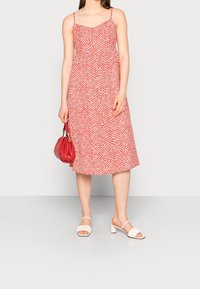 Rotes gemustertes Midi-Kleid mit dünnen Trägern, lockerem Schnitt und rundem Ausschnitt. Kombiniert mit weißen Blockabsatzsandalen und einer roten Handtasche.