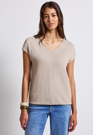 Beige textured top with a V-neck and short sleeves, paired with blue denim jeans. The fabric features a subtle raised pattern.
