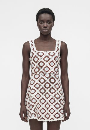 Woman wearing a sleeveless crochet dress with white and brown floral square patterns, standing against a plain light background.