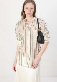 Light beige striped button-up shirt with black vertical lines, featuring a folded collar and single pocket, paired with a small black handbag.