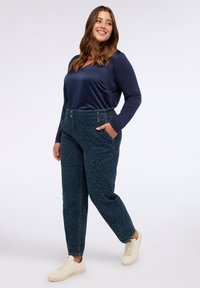 Navy-satin Langarmoberteil kombiniert mit türkisfarbenen, gemusterten, hochgeschnittenen Hosen mit Taschen und elastischem Bund, abgerundet mit hellfarbenen Sneakers.