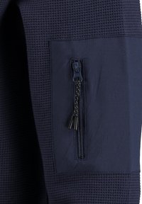 Donkerblauwe stof met een gestructureerd, wafelpatroon. Bevat een zijzak met een stevige ritssluiting en een decoratieve koordtrekker.