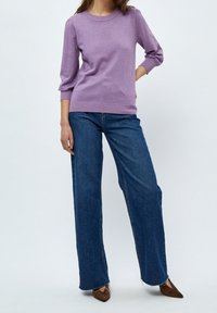 Person klädd i en lila långärmad tröja, vida blå jeans och bruna spetsiga skor, står med ena handen på höften.