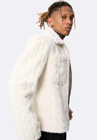 Homme avec des dreadlocks portant une veste en polaire texturée blanche, se tenant de côté contre un fond clair uni.