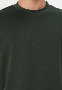 T-shirt vert foncé à col rond, avec une texture douce et des manches courtes, présentant un design simple et épuré sans motifs ni accents.