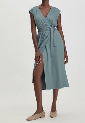 Donna che indossa un vestito incrociato verde salvia senza maniche con spacco laterale e cintura in tessuto coordinato, abbinato a scarpe basse beige.