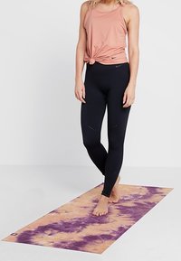 Tapis de yoga avec un motif tie-dye en mauve et pêche, assorti à un débardeur rose clair et un legging noir avec des détails perforés.