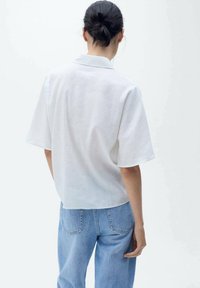 Camisa blanca de lino con un corte relajado, mangas cortas, un collar y la parte trasera ligeramente recortada, combinada con jeans de mezclilla azul claro.