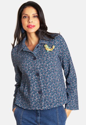 Frau trägt eine blaue, gemusterte Jacke mit Knöpfen und eine gelbe Brosche in Form eines Fisches, kombiniert mit blauen Jeans, mit den Händen in den Taschen, neutraler Hintergrund.