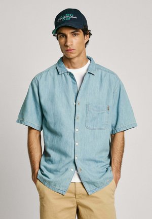 Lichtblauwe spijkerblouse met korte mouwen, knopsluiting en een enkele borstzak, gecombineerd met beige broeken en een marineblauw pet met groene accenten.