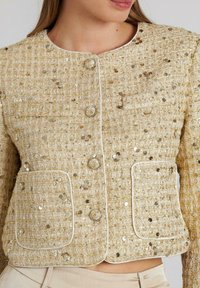 Goldfarbener, mit Pailletten besetzter Tweed-Jacke mit zwei vorderen Taschen, rundem Ausschnitt und Knopfverschluss. Strukturiertes Material mit kontrastierenden weißen Akzenten.