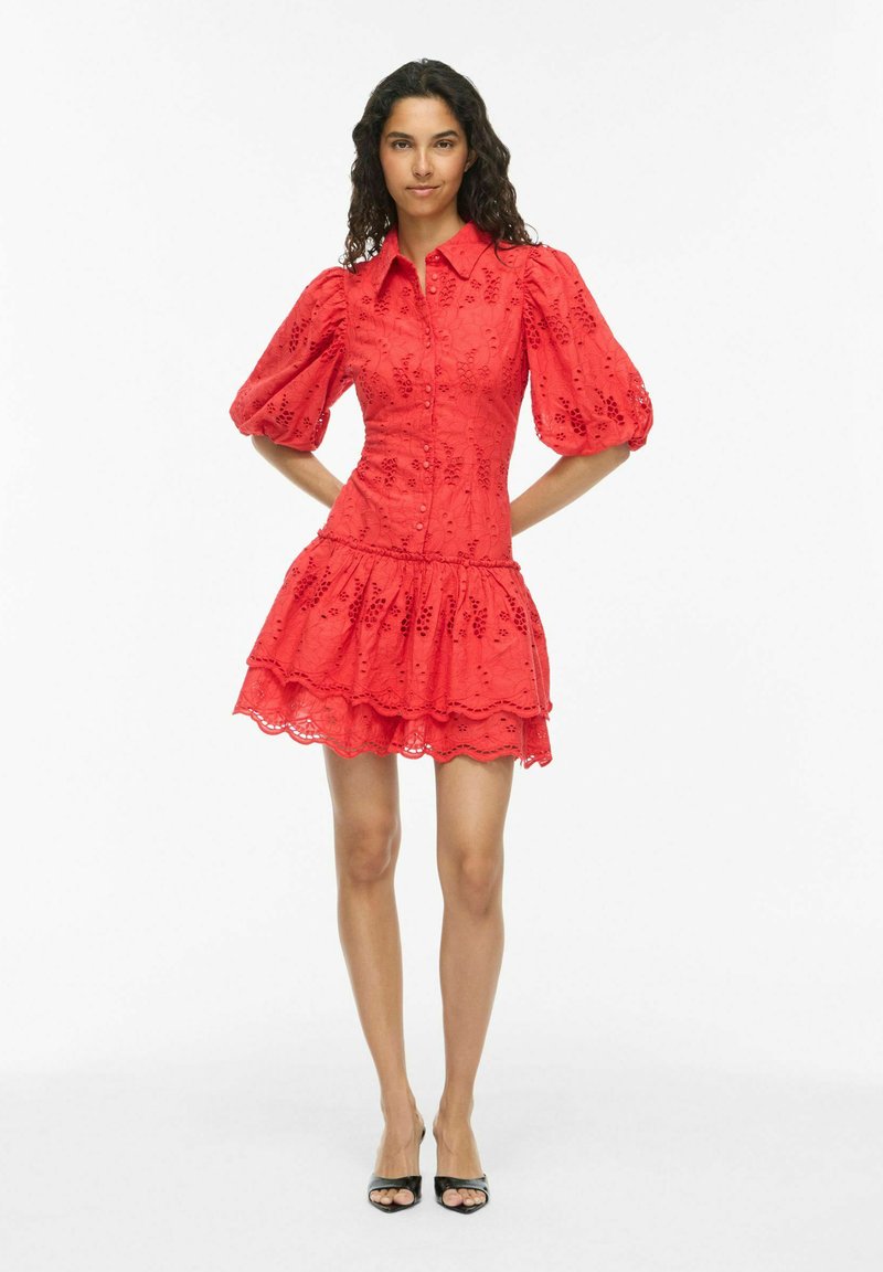 Femme portant une robe rouge en broderie anglaise avec manches bouffantes, une jupe à volants, et des talons noirs à bout ouvert, debout, mains derrière le dos.