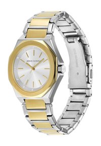 Nerezová ocelová hodinky s osmiúhelníkovým stříbrným a zlatým pouzdrem. Bílý ciferník má zlaté indexy a tři diamantové akcenty. Náramek ze stříbra a zlata.