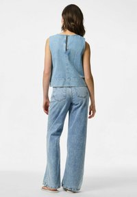 Traper prsluk s prilagođenim dizajnom, plave boje, bez rukava i s patentnim zatvaračem na leđima. Uparen s širokim plavim trapericama, oba imaju izblijedjeli izgled.