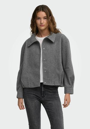 Jeune femme aux cheveux ondulés portant une veste en laine grise boutonnée sur une chemise blanche et un jean foncé sur un fond uni.
