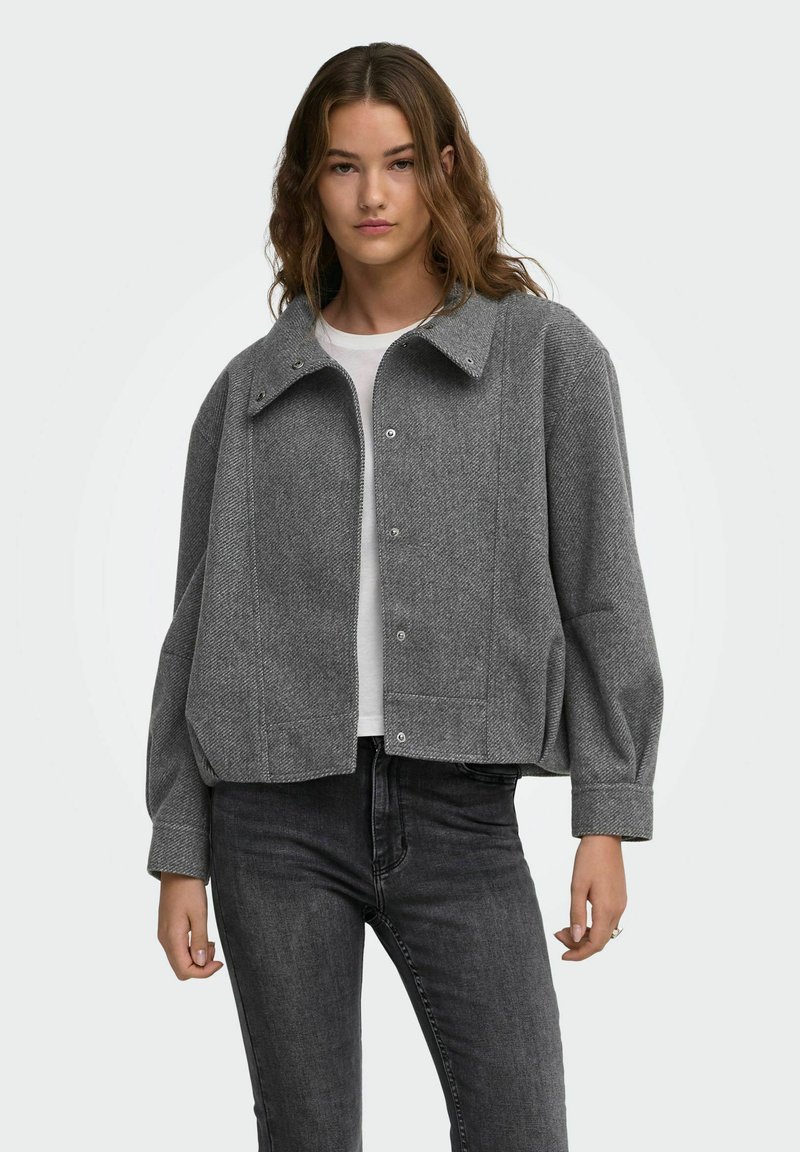 Jeune femme aux cheveux ondulés portant une veste en laine grise boutonnée sur une chemise blanche et un jean foncé sur un fond uni.