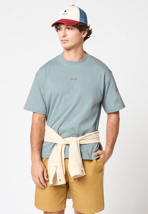Giovane uomo con maglietta azzurro chiaro, pantaloncini beige, giacca color crema legata in vita e cappellino da baseball rosso, blu e bianco che guarda di lato.