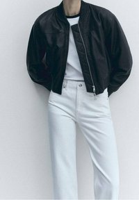 Chaqueta bomber de cuero negro con cuello acanalado, cierre de cremallera y detalles acolchados, combinada con vaqueros blancos de talle alto.