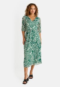 Robe midi verte à motif, avec un décolleté en V, des manches trois-quarts et une coupe fluide. Associée à des sandales noires à talons.