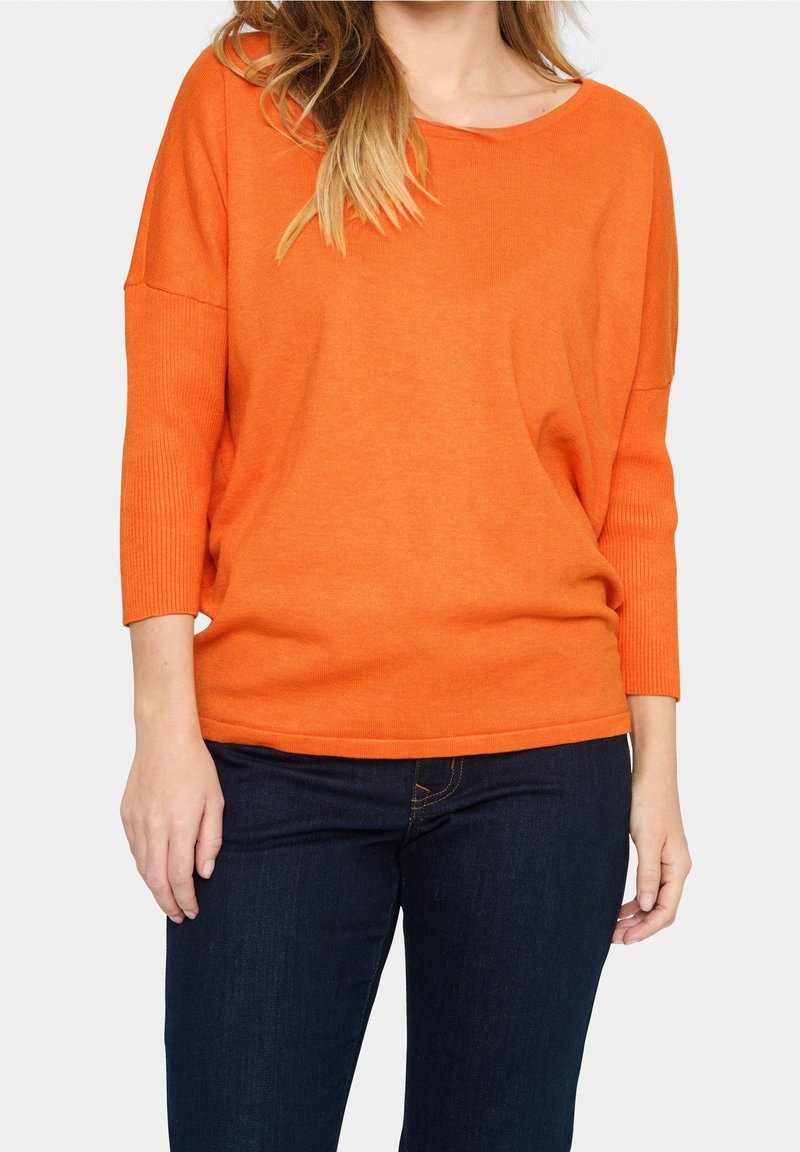 Femme portant un pull à manches longues orange vif avec un col bateau et un jean bleu foncé, debout devant un fond clair.