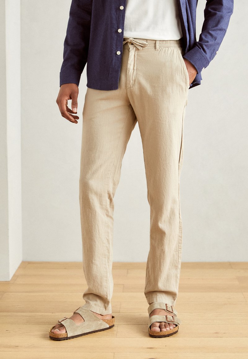 Beige linen trousers with a relaxed fit, featuring an elastic drawstring waistband. Paired with tan, open-toed sandals on a wooden floor.