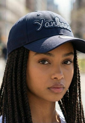 Young woman with long braided hair wearing a dark blue Yankees cap, looking slightly off camera in outdoor urban setting.