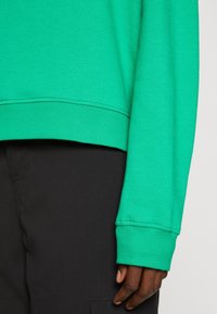 Sudadera verde con cuello redondo y puños acanalados, combinada con pantalones negros. La tela parece tener una textura suave.