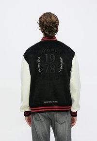 Άτομο με κυματιστά μαλλιά που φοράει μαύρο και άσπρο varsity μπουφάν με μανσέτες με κόκκινες ρίγες και κείμενο «1978» και «Broke Since Milano» στην πλάτη.