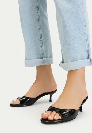 Pies con vaqueros azul claro remangados y zuecos de tacón bajo tipo gatito, abiertos en la punta, de charol negro, sobre un fondo blanco.