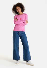 Lächelnde Frau mit lockigem Haar steht mit verschränkten Armen, trägt ein pinkes Langarmshirt mit einem roten Herzen, weitgeschnittene blaue Jeans und weiße Turnschuhe.