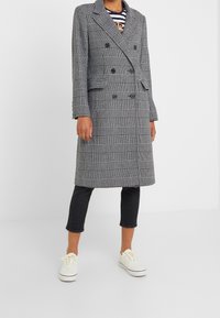 Černobílý kabát s houndstooth vzorem, dvouřadový, s velkými klopami, předními kapsami a přímým střihem. V kombinaci s tmavými džíny a bílými teniskami.