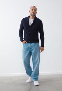 Navy knitted cardigan with a shawl collar, five buttons, and two front pockets, paired with light blue loose-fitting jeans and white sneakers.