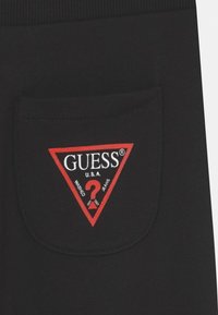 Schwarzer Stoff mit einem dreieckigen roten GUESS-Logo, das weiße Schrift und ein Fragezeichen zeigt, auf einer Gesäßtasche gestickt.